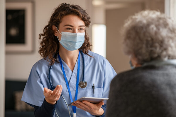 A nurse wears a safety protective mask while talking with a senior patient.